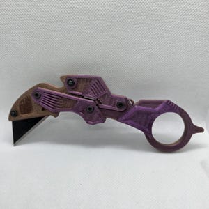 May include: A 3D-printed utility knife with a curved blade and a finger ring. The knife features a brown and purple color scheme with visible screws and geometric designs. The blade is black.
