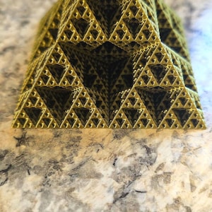 May include: A geometric, golden-colored fractal pyramid. The pyramid is constructed from repeating triangular patterns, creating a complex, open structure. The object is displayed on a speckled, light-colored surface.