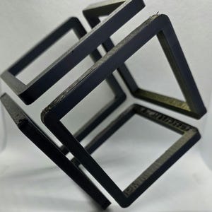 May include: A black, three-dimensional geometric sculpture featuring interconnected square frames, creating an optical illusion. The artwork is set against a white background, highlighting the dark color and intricate design.