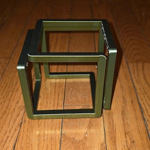 May include: A green metal cube-shaped object with a hollow interior. The cube is made of thin, rectangular bars joined at right angles. The object is resting on a wooden surface.