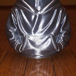 Può includere: Una felpa con cappuccio argentata metallizzata con tasca a marsupio e cappuccio. Il materiale lucido riflette la luce, creando riflessi e ombre. La felpa con cappuccio è esposta su una superficie di legno.