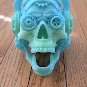 May include: A teal and green skull sculpture with headphones and goggles. The skull has an open mouth and is resting on a wooden surface. The sculpture is a unique decorative item.