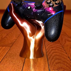Könnte beinhalten: Ein schwarzer und galaxie-themenbezogener Videospiel-Controller ruht auf einem bronzefarbenen, handförmigen Ständer. Der Controller hat schwarze Joysticks und Tasten mit blauen Akzenten. Der Ständer steht auf einer Holzoberfläche.