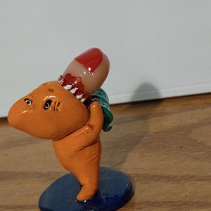 May include: A quirky orange shark figure, biting a finger. The finger has red nail polish and a bite mark. The shark stands on a blue base and has green fins. The figurine is on a wooden surface.