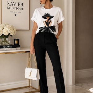 Könnte beinhalten: Ein weißes kurzärmeliges T-Shirt mit einer grafischen Illustration einer Figur in schwarzem Hut und Kleid, kombiniert mit einer schwarzen, maßgeschneiderten Hose, einer weißen, strukturierten Handtasche mit Goldkette und weißen Pumps.