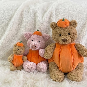 Könnte beinhalten: Ein kleiner brauner Teddybär, ein rosa Schweinchen-Plüschtier und ein großer brauner Teddybär, die jeweils ein orangefarbenes Kürbiskostüm mit einem kleinen grünen Stiel tragen. Die Plüschtiere haben weiches, strukturiertes Fell.