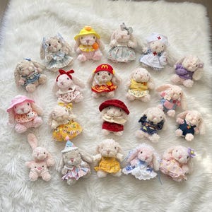 May include: A collection of twenty small plush rabbit dolls, each wearing different miniature outfits including dresses, hats, overalls, and uniforms. The rabbits have light-coloured fur and are arranged in a group on a white textured surface.