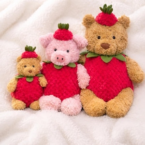 May include: Three plush toys consisting of two tan bears and one pink pig, each wearing a red strawberry-themed outfit with green leaf accents on the head and neck. The toys feature textured red bodies, dark eyes, and soft, fuzzy fabric construction.