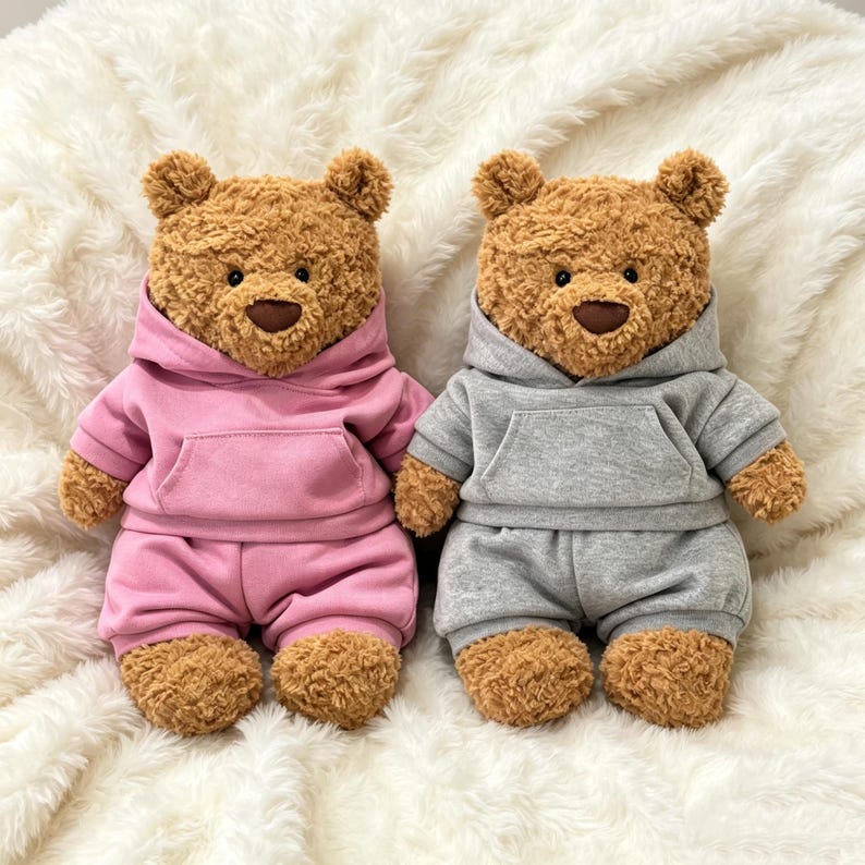 May include: Two plush teddy bears with brown curly fur and dark brown noses. One bear wears a pink hooded sweatshirt and matching shorts, while the other bear wears a gray hooded sweatshirt and matching shorts. Both bears have a front pocket.