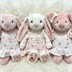 Puede incluir: Tres peluches formados por dos conejos y un cerdo, cada uno con un vestido blanco con estampado floral y una etiqueta de Hello Kitty. Los conejos tienen orejas largas con forro floral y el cerdo tiene un lazo blanco en la oreja.
