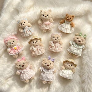 May include: A collection of ten small plush teddy bears arranged in a group. Each bear features soft, light brown fur and is dressed in a unique outfit, including various patterned dresses, hats, bows, and lace details in pastel colours and white.