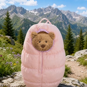 Op de afbeelding: Een bruine pluche teddybeer in een lichtroze, gewatteerde slaapzak-achtige pod. De pod heeft een afgeronde bovenkant met een klein handvat en een ronde opening die het gezicht van de beer onthult, omlijst door een zachte, lichtpaarse voering.