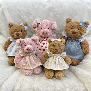 Puede incluir: Cinco peluches compuestos por tres osos de peluche marrones y dos cerditos rosas. Cada juguete lleva un vestido de tela estampado con ribete de encaje y un pequeño lazo en la cabeza. Los vestidos presentan estampados de flores, fresas y margaritas.