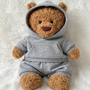 May include: A brown teddy bear with textured fur, black eyes, and a dark brown nose. The bear is wearing a gray hooded sweatshirt with a front kangaroo pocket and matching gray sweatpants that cover the lower body and legs.