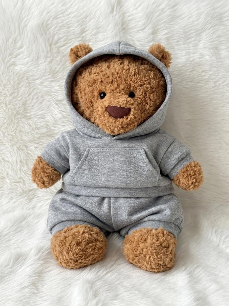 May include: A brown teddy bear with textured fur, black eyes, and a dark brown nose. The bear is wearing a gray hooded sweatshirt with a front kangaroo pocket and matching gray sweatpants that cover the lower body and legs.