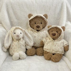 Puede incluir: Un conejo de peluche blanco y dos osos de peluche marrones, cada uno con una chaqueta acolchada de color crema con cremallera, un delicado estampado floral rosa y capucha. Los peluches tienen pelaje suave, ojos oscuros y narices marrones.