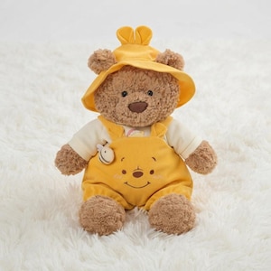 May include: A plush brown teddy bear wearing a yellow bucket hat with small ears and yellow overalls over a cream-coloured shirt. The overalls feature a small embroidered bee on the chest and a stitched face of a bear on the front pocket.