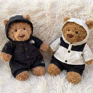 May include: Two brown plush teddy bears wearing hooded jackets and pants. The bear on the left wears an all-black outfit with a white logo on the chest. The bear on the right wears a white and black color-blocked jacket with black pants.