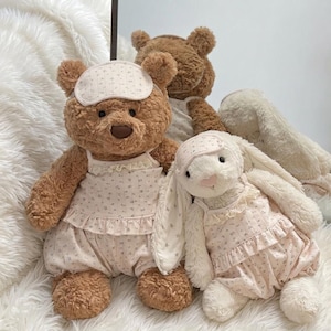 May include: A brown plush bear and a white plush rabbit wearing matching cream-colored floral print camisole tops, ruffled bloomers, and sleep masks. The bear has dark brown fur and a brown nose, while the rabbit has long ears and a pink nose.