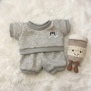 May include: A heather grey short-sleeved sweatshirt and matching shorts set featuring a small embroidered black cat graphic on the chest. Beside the clothing is a plush, tan-coloured coffee cup toy with a smiling face and dangling brown legs.