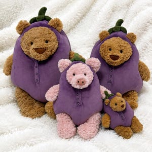 May include: Three plush toys consisting of two brown bears and one pink pig, each wearing a purple aubergine-shaped onesie with a green stem on the head. The onesies feature a button-down front detail. The toys vary in size from large to small.
