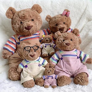 May include: A collection of six brown plush teddy bears of varying sizes. The bears wear striped shirts and corduroy overalls in shades of tan, pink, and cream. One bear wears black-rimmed glasses, and another has a small red and white checkered bow.