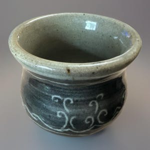 Könnte beinhalten: Ein handgefertigter Keramiktopf mit breitem, abgerundetem Rand und dunkelgrauem Körper. Der Topf hat ein weißes, wellenartiges Design um den Boden. Der Innenraum und der Rand sind ein helleres, gesprenkeltes Grau. Ideal zum Halten kleiner Gegenstände.