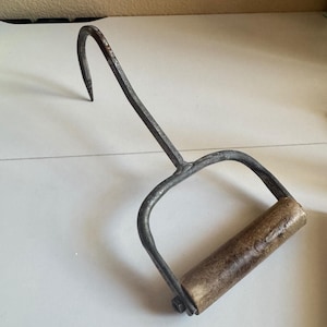 May include: A hand tool featuring a curved, pointed metal hook attached to a metal frame with a cylindrical wooden handle. The metal components have a dark, textured finish, while the wooden handle shows a light brown, worn grain.