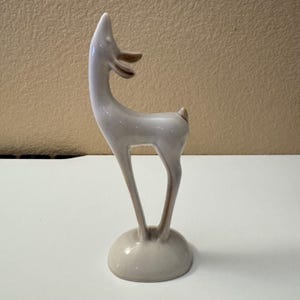May include: A stylized ceramic deer figurine with a glossy white finish and light brown accents on the ears and tail. The deer stands on a rounded base with an elongated neck and slender legs, featuring a smooth, reflective surface throughout.