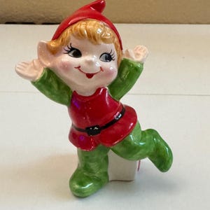 Puede incluir: Una figura de elfo de cerámica con acabado brillante, que lleva un gorro rojo, una túnica roja con cinturón negro y pantalones verdes. El elfo tiene cabello rubio, orejas puntiagudas y un rostro expresivo con ojos grandes y brazos alzados.