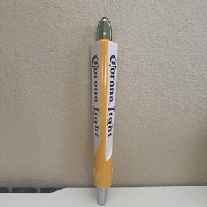 May include: A vertical Corona Light beer tap handle featuring a yellow base and white sides with the black text Corona Light printed on both sides. The top of the handle is shaped like a green lime wedge, and the bottom has a silver metal connector.