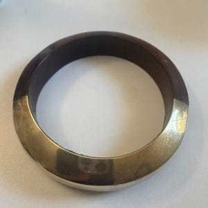 May include: A circular bangle bracelet featuring a two-tone design. One half of the bangle is crafted from a smooth, dark brown material, while the other half is made of a polished, reflective metallic gold-toned material with a subtle hammered texture.