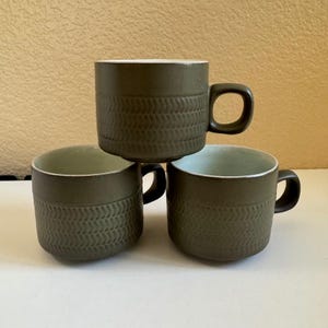 May include: Three olive green ceramic mugs with a matte finish and a textured herringbone pattern around the lower half. Each mug features a rounded handle and a smooth, light-colored interior. The mugs are arranged in a stack of three.