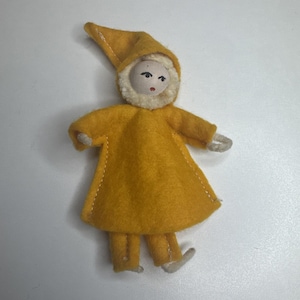 Puede incluir: Una pequeña muñeca hecha de fieltro amarillo, con una capucha puntiaguda y un borde de pelusa color crema alrededor de la cara. La muñeca tiene una cabeza pálida y redondeada con ojos negros pintados y boca roja, vistiendo una túnica amarilla.