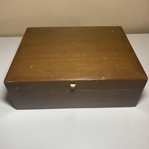 May include: A rectangular wooden box with a medium brown finish and a smooth, flat lid. The box features a small, centred gold-toned metal latch on the front edge. The wood surface shows minor signs of wear and light surface scratches.