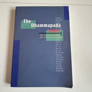 May include: A blue book titled The Dhammapada by Narada, featuring a list of chapters including Yamaka Vagga and Bhikkhu Vagga. The cover includes green rectangular accents, white text, and the subtitle PALI text & translation with stories in brief & notes.
