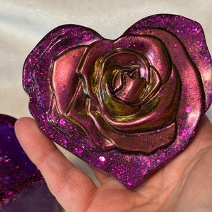 May include: A heart-shaped decorative piece with a rose design. The piece is a deep purple color with glitter and iridescent accents. The rose has a metallic finish with shades of gold and pink.