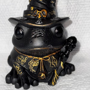 Può includere: Una statuetta di rana nera con cappello e veste da strega, con dettagli dorati. La rana tiene in mano un bastone, e il cappello e la veste sono adornati con intricati motivi dorati. La statuetta è posta su uno sfondo bianco.