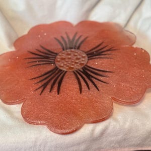 May include: A decorative, flower-shaped resin tray in shades of coral with glitter accents. The center features a dark brown floral design. The tray is resting on a white surface.