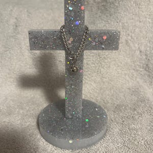 Puede incluir: Una cruz decorativa hecha de resina gris con purpurina incrustada. Una cadena plateada con un pequeño dije cuelga de la cruz. La cruz está montada sobre una base circular.