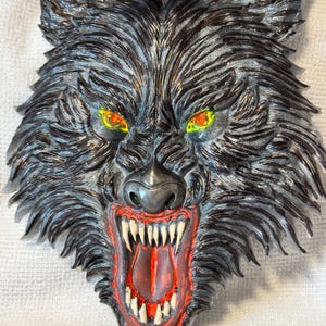 May include: A detailed, three-dimensional wolf head sculpture with an open mouth, revealing sharp white teeth and a red interior. The wolf's fur is primarily black with silver highlights, and its eyes are yellow and orange. The sculpture has a menacing appearance.