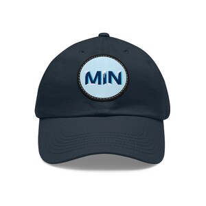May include: Navy blue baseball cap with a circular patch featuring the letters "MIN" in white and blue, set against a light blue background. The patch is outlined in black and white, and the cap has a curved brim.