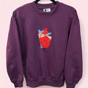 May include: A deep purple sweatshirt with a crew neck and long sleeves. The front features a stitched anatomical heart design in red, blue, and plaid fabric. The tag reads "SPIRIT".