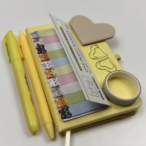 Op de afbeelding: Een verzameling gele briefpapierartikelen, waaronder een notitieboek, pennen, een markeerstift, plaknotities met kattenontwerpen, een hartvormige leren patch, paperclips en washitape. Het notitieboek heeft een lint.