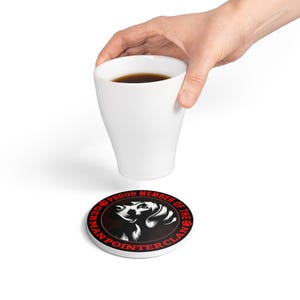 May include: White ceramic coaster with a black and white German Pointer dog illustration and the text "PROUD MEMBER OF THE GERMAN POINTER CLAN" in red. A white coffee cup is held above the coaster.