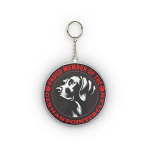 Może przedstawiać: Okrągły brelok do kluczy ze srebrną obwódką i grafiką psa. Brelok przedstawia czarno-białą ilustrację psa rasy Wyżeł Niemiecki. Tekst "PROUD MEMBER OF THE GERMAN POINTER CLAN" jest wydrukowany na czerwono na obrzeżu.