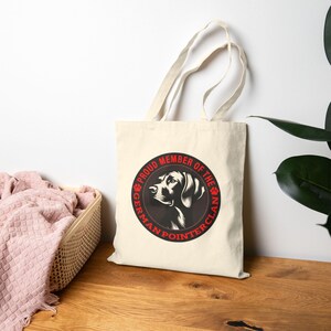 May include: A cream-colored tote bag featuring a circular graphic of a German Pointer dog with the text "PROUD MEMBER OF THE GERMAN POINTER CLAN" in red. The bag has two handles and is made of a canvas-like material.