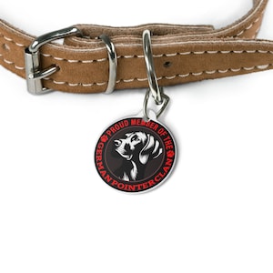 Op de afbeelding: Een bruine leren hondenhalsband met een zilveren gesp en een ronde hondenpenning. De hondenpenning toont een zwart-wit illustratie van een Duitse Staande Hond met de rode tekst "PROUD MEMBER OF THE GERMAN POINTER CLAN".