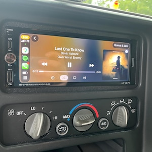 May include: A black car stereo head unit with a touchscreen display showing a music interface. The screen displays the song Last One To Know by Gavin Adcock, along with navigation icons, signal strength, and time. Below are three circular climate control knobs.