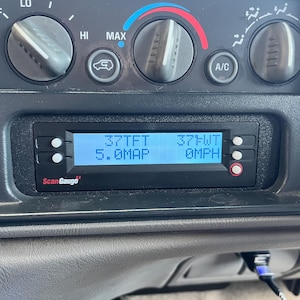 May include: A black ScanGauge device is installed in a vehicle's dashboard. The digital display shows readings in white text against a blue background. The device is surrounded by climate control knobs and vents.